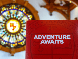 ChatGPT said: Red booklet with the words “Adventure Awaits” held in front of a colorful stained-glass window at Saint Lawrence University.