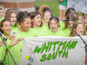 A group of students wearing bright green shirts cheer and hold a painted banner that reads “Whitman South” during an outdoor event at Saint Lawrence University.