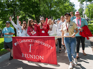 A group of smiling people walk outdoors holding a red banner that reads “Saint Lawrence University 1st Reunion.”
