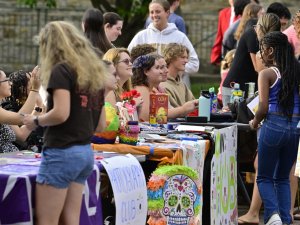 Students gather around tables at an outdoor student club fair at Saint Lawrence University, chatting and signing up for activities. Tables are decorated with colorful signs, posters, and props, including a piñata-style skull and a "Photography Club" banner.