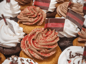 A tray of cupcakes topped with white or chocolate frosting, some with red sprinkles and small red-and-white striped flags.