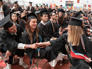 Graduating students dressed in caps and gowns sit in rows, smiling and interacting with one another. Two students in the foreground shake hands, holding their diplomas and sharing a joyful moment. The space is decorated with international flags, emphasizing a celebratory and inclusive atmosphere.