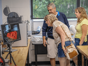 Three people lean in to examine a computer monitor displaying a bright orange 3D-printed lattice structure inside a lab with workbenches and equipment.