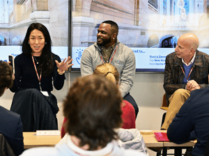 Three panelists sit at the front of a room during a discussion, speaking to an audience seated in rows. One person gestures while talking, and the others listen and smile. A screen behind them displays presentation content in a bright, modern space.