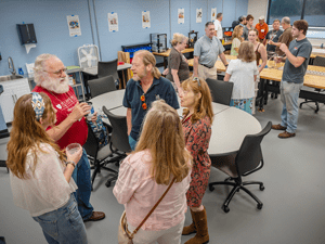 Professors, parents, and college students stand around round tables in a classroom, talking in small groups. A professor with a white beard sits in a wheelchair holding a water bottle while speaking with three college students. Other professors, parents, and college students chat in the background near tables, chairs, and posters on the walls.