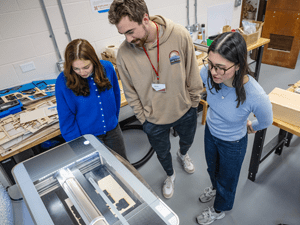 Three people stand around a laser cutter in a makerspace, looking down at a project in progress, with wood pieces and tools arranged on nearby tables.