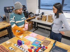 Two people stand at a workbench in a makerspace, discussing colorful 3D-printed prototypes, molds, and sample pieces spread out on the table.