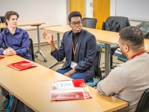 Two students meet with an alum at a table during a networking conversation. One student gestures while speaking as the others listen, with red folders and papers on the table. The discussion takes place in a classroom at Saint Lawrence University.
