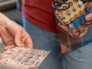 Close-up of hands holding a small etched circuit board and a wooden mechanical assembly with gears, bolts, and colorful wires attached.