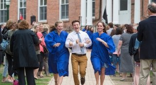 Three Saint Lawrence students run down a side walk and laugh  as a crowd watches on. Two students are wearing vibrant blue robes. 
