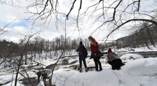 A wintery outdoor scene. Three students stand on a snow-covered trail overlooking a waterfall.