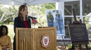 President Kate Morris stands behind a podium under a white tent and next to the "Jeffrey Campbell House" dedication plaque.