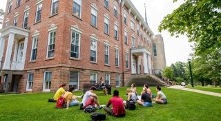 Professor Sarah Gates sits on a bench outside of Richardson Hall teaching a class to ten Saint Lawrence University students on a sunny day. 
