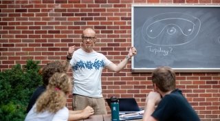 Professor Dan Look, standing in front of a picnic table outside, holds his hands up to explain a concept while teaching a class to three Saint Lawrence students. Behind Dan, there is a black chalkboard affixed to a brick wall. 