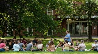 Laura Mills Smith stands teaching a psychology class outside on a bright, sunny day. Several Saint Lawrence University students sit on the grass with notebooks.  
