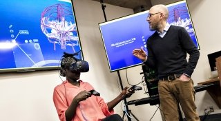 A student sits as they explore a virtual reality through a headset they are wearing as a faculty member describes the experience.