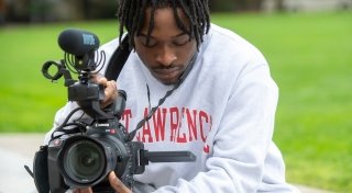 Kaleb Davis, wearing a Saint Lawrence sweatshirt, looks at the digital display of a large video camera. 