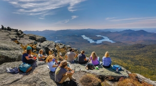 A group of students wearing hiking gear sit on the rocky face of Whiteface Mountain, gazing out at the Adirondack hills, peaks, and lakes beyond. It's a beautiful, clear day.