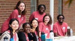 Seven Saint Lawrence University students, wearing brick red t-shirts and name tags, smile as they sit at a long table outside on a sunny day. 
