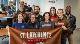 A group of Saint Lawrence University alumni, wearing Saint Lawrence sweatshirts and t-shirts, stand in a group and hold up the Saint Lawrence Saints brown flag.