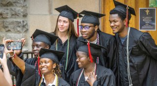 Six Saint Lawrence University graduates in caps and gowns pose for a selfie outside a stone building, smiling and celebrating together.