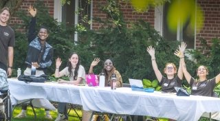 A group of smiling Saint Lawrence University students sit and stand behind a long table outdoors, waving and raising their hands. The table is covered with a white cloth and has laptops, water bottles, and notebooks on it. Green shrubs and a brick building are in the background.