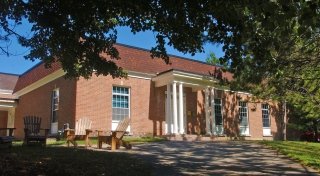 Gaines Hall at Saint Lawrence University is a red-brick building with white columns and large windows. The front lawn includes wooden chairs for outdoor seating under the shade of nearby trees. The building has a classic look and is surrounded by green space. 