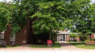 Lee Hall at Saint Lawrence University is a red-brick building surrounded by trees and green space. A path leads to a glass entryway, and outdoor seating is available with benches and Adirondack chairs. A red sign with white text marks the building’s name.