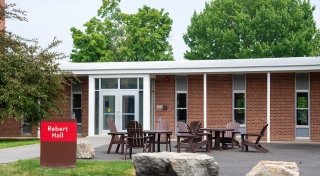 Exterior of Rebert Hall with a red-brick facade, glass entrance doors, a red sign, and outdoor seating with Adirondack chairs and tables.