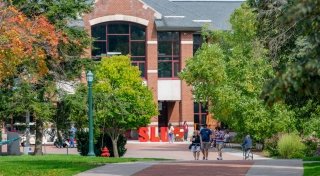 People walk and bike along a tree-lined path toward a red brick campus building with large windows at Saint Lawrence University, with large red "SLU" letters in front.