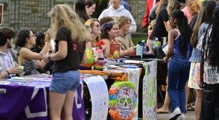 Students gather around tables at an outdoor student club fair at Saint Lawrence University, chatting and signing up for activities. Tables are decorated with colorful signs, posters, and props, including a piñata-style skull and a "Photography Club" banner.