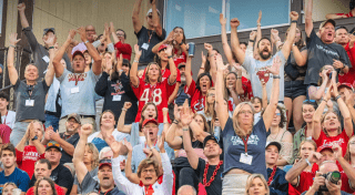 A crowd of Saints fans raise their hands and cheer while watching a football game. 