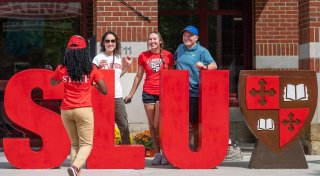 Four people smile and pose behind large red "SLU" letters and a Saint Lawrence University shield outside a brick building, while a staff member in a red shirt and cap faces them.