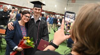 A graduate in cap and gown smiles with a family member holding flowers, while another person takes their photo with a phone during a Saint Lawrence University commencement ceremony.