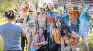 A group of Saint Lawrence University students smile and pose outdoors during a color run, covered in bright powder paint while one person takes a photo. The group stands on a grassy campus area with trees and a building in the background, showing a fun and energetic campus event.