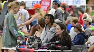 Students sit and stand at a crowded outdoor table during a campus event at Saint Lawrence University, chatting and smiling while surrounded by gear and club materials.
