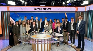 A large group of students and two adults pose together in a CBS News studio behind a round glass desk. The set features bright studio lights, a blue world map backdrop, and a banner reading “CBS News” above them. Most people wear business casual clothing and name badges, suggesting a group visit or tour..