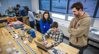 Five students work together in a campus makerspace, gathered around a wooden table with laptops, wires, and electronic components. One student types on a laptop while others watch and discuss, and more students use equipment in the background. Large windows line the wall, bringing natural light into the lab setting.