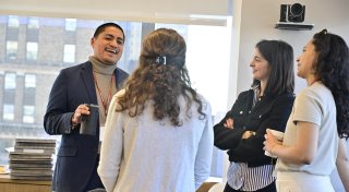 Four people stand in a small group near large windows, talking and smiling during a casual networking moment. One person holds a travel mug while two others hold coffee cups, and a stack of notebooks sits on a nearby table. A city building is visible through the window behind them.