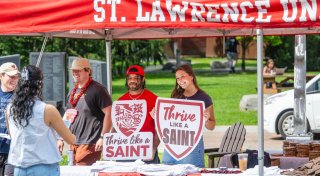 Four individuals stand under a red canopy with "ST. LAWRENCE UN" visible, holding signs that read "Thrive like a SAINT" and featuring a shield emblem with "1856," while others interact nearby.
