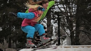 Two students wearing neon snow pants and jackets ski off of a jump together.