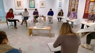At least 10 students sit in a large circle with large pads of paper in an art gallery as they draw a statue in the middle.