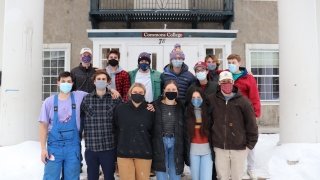 A group of twelve students stand in two rows on the snowy front steps of their residence. 