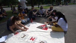 A group of students sit on the sidewalk decorating a flag to represent their First Year Program college. 