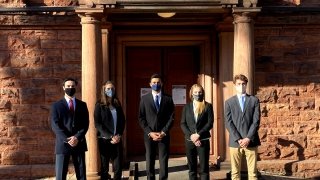 Five students in business clothing stand in front of a sandstone building.