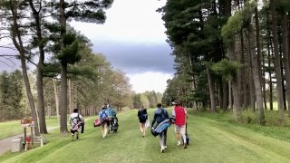 Six students walk with golf bags on some of their shoulders on the golf course.