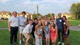 A group of students thirteen students gather and pose for a photo on a lawn on a summer day.