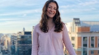 Lily MacCachran wearing a light pink blouse and standing on a balcony overlooking tall buildings in the background.