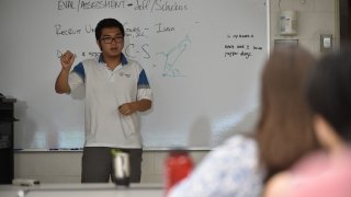 A student stands at a whiteboard at the front of the class explaining a math formula to their audience.