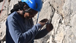 A student wearing a helmet and a harness, prepares to climb the rock face in front of them.